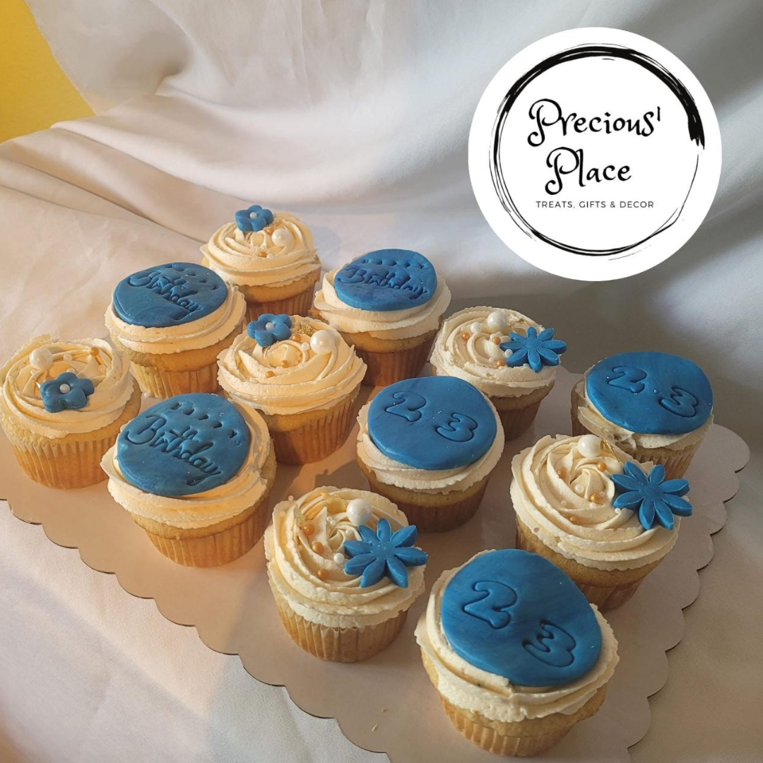 A dozen decorative cupcakes with blue fondant decorations, including 'Birthday' and '23' inscriptions, accompanied by floral accents.